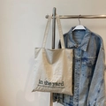 Große beige Segeltuch-Tasche mit Grafikdruck an einem Metallständer neben einer Jeansjacke - Cremeweiß