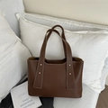 Bolso tote marrón oscuro con costuras blancas y remaches dorados en un sofá - Marrón