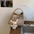 Detail der beige Eimer-Tasche mit Litschimuster-Leder, Metallschnalle und Kaninchenanhänger - Khaki Mit Anhänger