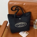 Minimalist black canvas tote bag with white oval logo print and silver snap button closure - Black