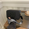 Chic black camera bag with SM logo and geometric wide strap on a cafe table - Black