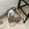 Detail of taupe leather bucket bag with cute white plush rabbit charm on a cozy bed - With Charm / Tan