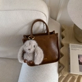 Close-up of brown faux leather handbag with a fluffy rabbit charm on a cream sofa - Brown with Pendant
