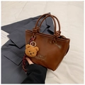 Braune Handtasche aus Leder mit Bärenanhänger und Text auf weißem Stoffhintergrund - Braun