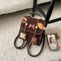 Detalle de una mini mochila de ante marrón con adornos de peluche y un lazo rojo - Marrón