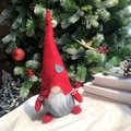 Red plush Christmas gnome with gray heart patch and striped sleeves on holiday tree background - Male Style