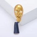 Artistic gold and blue thinker face mask brooch shown on a white geometric stand - Gold
