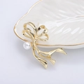 Elegante broche de lazo dorado con perla blanca en una bandeja de cerámica minimalista - Dorado