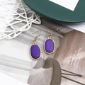 Detail view of golden hook earrings featuring purple oval resin stones with metallic borders - Purple