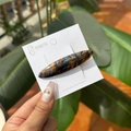 Détail macro d'une barrette en acétate écaille de tortue avec lettrage doré sur fond de feuilles - Pince Bec de Canard Ovale Étiquette Dorée / Marbre Peinture à l'Huile Bleu Brun