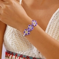Primer plano de una pulsera tejida bohemia con flores de plumeria moradas y cristales finos - Púrpura