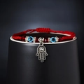Red braided evil eye bracelet with Hamsa Hand charm and blue beads on a black ceramic display - Hamsa Style