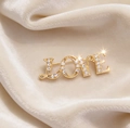 Dettaglio macro della spilla LOVE dorata con strass bianchi su velluto beige - Stile Lettera LOVE (6210403351049) / Oro