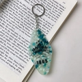Détail d'un porte-clés en acrylique en forme de losange avec motifs outdoor sur un livre - Boucle Dorée / Cyan