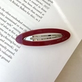 Detail of a dark red oval acetate hair clip with AMY BEAR logo resting on book pages - Red