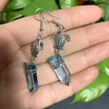 Detail of silver turtle earrings with sea blue crystal stones held in hand against foliage - Light Blue