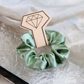 Lustrous sage green satin hair scrunchie displayed on a diamond graphic card holder - Green