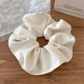 Detalle de coletero scrunchie en tela texturizada crema con etiqueta sobre fondo decorativo - Blanco Roto
