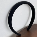 Close-up of a thick black padded satin headband leaning against a white textured wall - Black