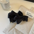 Détail d'une pince à cheveux à nœud en satin noir avec un petit cœur doré sur du beige - Noir