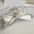 Dettaglio del fermaglio trasparente con fiocco crema, perle e strass su sfondo neutro - Bianco con Fiocco