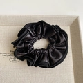 Close-up of black pleated faux leather scrunchie with detailed stitching in a beige box - Black Hair Tie