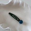 Detailed view of dark green metal drop hair clip with blue bead on white ceramic plate - Green