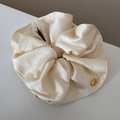Primer plano detallado de un coletero de satén crema con una etiqueta metálica dorada - Scrunchie / Coletero Blanco Crema Etiqueta Dorada