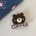 Detailansicht einer braunen Bären-Anstecknadel mit rosa COFFEE-Schriftzug auf Denim-Stoff - Dunkelkaffee Bärenkopf
