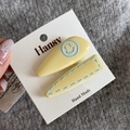 Close-up of yellow geometric hair clips with blue smiley and stitching on a grey background - Yellow