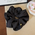 Detailed close-up of a black satin scrunchie featuring a golden metal logo on textured mat - Black