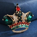 Elegante spilla a corona dorata con gemme verdi e cristalli su un tessuto blu scuro - Corona Verde Smeraldo