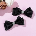 Dettaglio di due fermagli a fiocco in raso nero con strass a cuore su uno sfondo rosa delicato - Nero