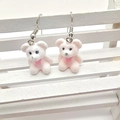 Detail view of cute pink flocked bear earrings with bows on a miniature white bench - Pink Bow Bear