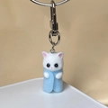 Detailed close-up of a cute flocked white kitten keychain wearing light blue felt clothes - Blue