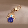 Goldener Hamsa-Hand Schlüsselanhänger mit blauem Auge-Anhänger auf braunem Stoffhintergrund - Hohle Handfläche / Gold
