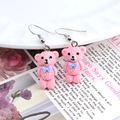 Gros plan de boucles d'oreilles ours rose avec nœud bleu et crochets argentés sur fleurs - Ours Rose