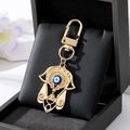 Gold Hamsa Hand keychain with blue Evil Eye charm on a black jewelry cushion background - Gold Hollow Hamsa