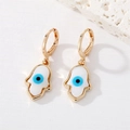 Golden Hamsa hand earrings with blue evil eye detail on a minimalist stone background - Hamsa Eye