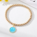 Golden beaded bracelet with sky blue enamel smiley face charm on a white stone background - Blue Smiley Face Bracelet