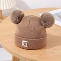 Detail of warm brown knit baby hat with dual pom-poms and fabric label on a wood table - Coffee