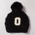 Close-up of black knitted baby hat with large fur pompom and gold sequin letter O patch - Black