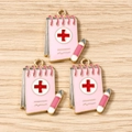 Macro detail of three pink medical notebook charms with red cross and golden metal edges - Pink