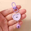 Detalle de llavero de corazón morado con cara sonriente, cierre de mosquetón y cascabel - Forma de Corazón / Morado