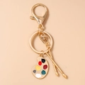 Artistic keychain detail with gold palette and paintbrushes charm on beige backdrop - Gold