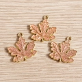 Detail von Ahornblatt-Anhängern aus Emaille in Pink und Gold auf hellem Holzgrund - Rosa