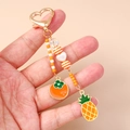 Nahaufnahme von goldenem Herz-Schlüsselanhänger mit Emaille-Ananas und Kaki-Anhängern - Orange Ananas