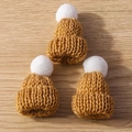 Detailed close-up of three camel knit mini hats with white pompoms on a wood background - 10 Pieces/Pack / Brown