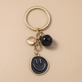 Goudkleurige sleutelhanger met zwarte smiley, hartje en belletje op een beige basis - Zwarte Smiley Face