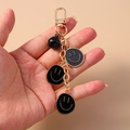 Handheld weergave van een zwarte smiley sleutelhanger met gouden ketting en belletje - Zwart